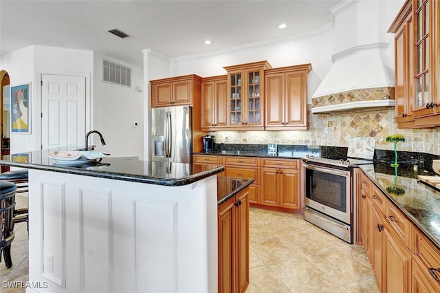 kitchen featuring dark stone counters, ornamental molding, an island with sink, custom range hood, and stainless steel appliances