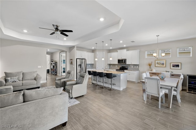 living room with ceiling fan with notable chandelier, a raised ceiling, beverage cooler, and sink