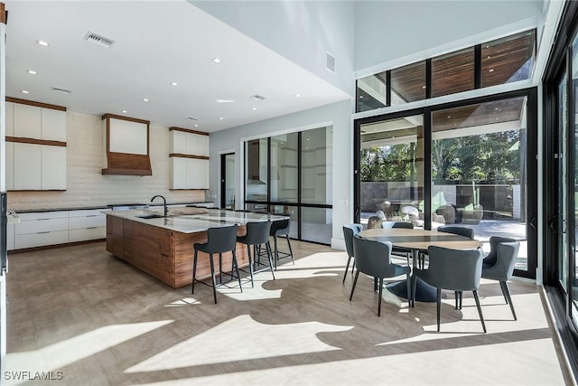 kitchen featuring backsplash, custom exhaust hood, sink, a center island with sink, and white cabinets