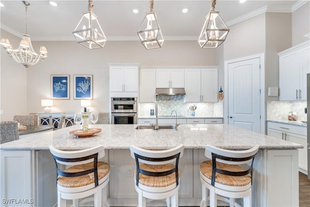 kitchen featuring white cabinetry, stainless steel double oven, a large island with sink, decorative light fixtures, and a kitchen bar