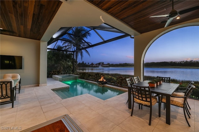 pool at dusk featuring glass enclosure, a water view, ceiling fan, an in ground hot tub, and a patio area