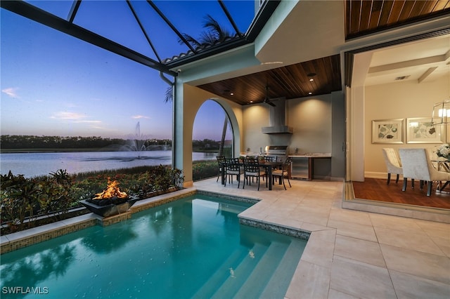 pool at dusk with glass enclosure, ceiling fan, a grill, a patio area, and a water view