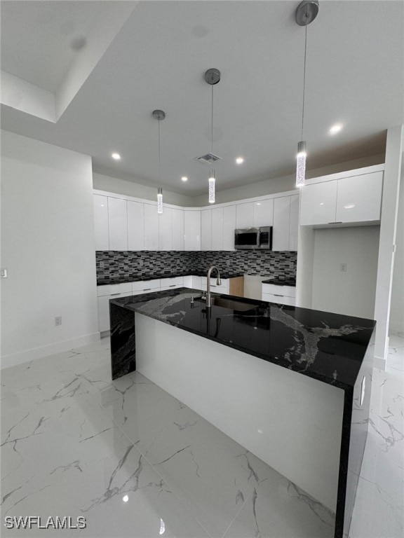 kitchen with white cabinetry, backsplash, dark stone counters, decorative light fixtures, and a kitchen island with sink