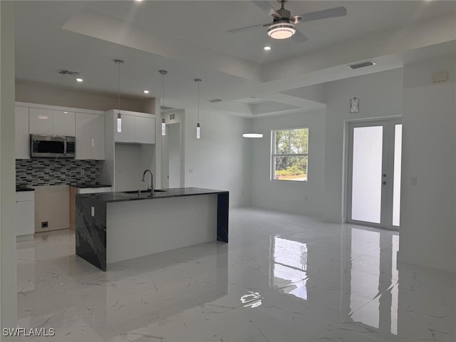 kitchen with backsplash, a kitchen island with sink, ceiling fan, white cabinets, and hanging light fixtures