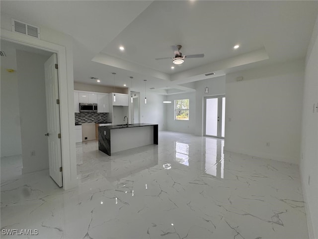 kitchen featuring ceiling fan, backsplash, a tray ceiling, a center island with sink, and white cabinets