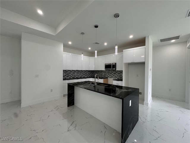 kitchen featuring white cabinetry, tasteful backsplash, dark stone counters, pendant lighting, and a center island with sink