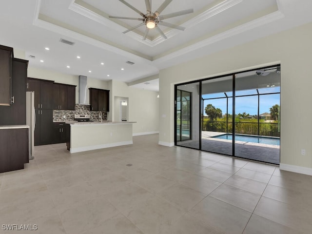 kitchen featuring ceiling fan, wall chimney exhaust hood, backsplash, a tray ceiling, and a kitchen island with sink