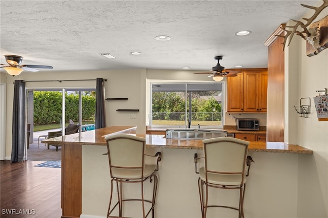 kitchen with brown cabinetry, a breakfast bar area, stainless steel microwave, dark wood-type flooring, and light stone counters