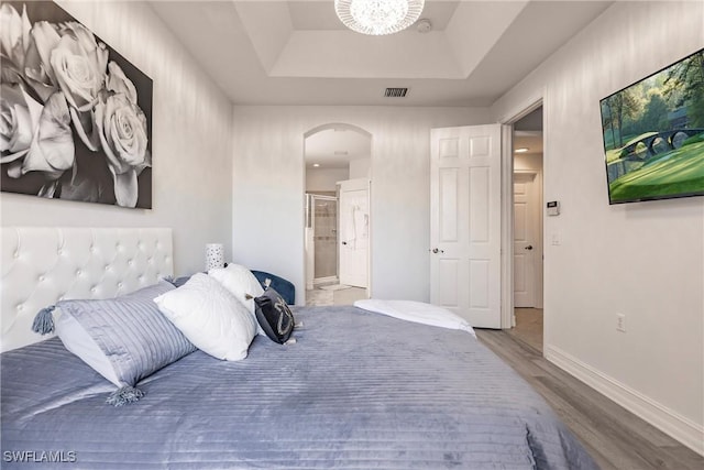 bedroom featuring a tray ceiling, ensuite bathroom, wood-type flooring, and a notable chandelier