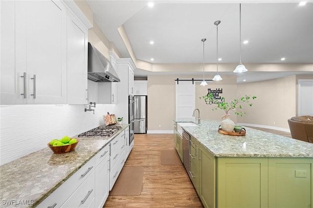 kitchen featuring wall chimney range hood, a barn door, decorative light fixtures, a center island with sink, and green cabinetry