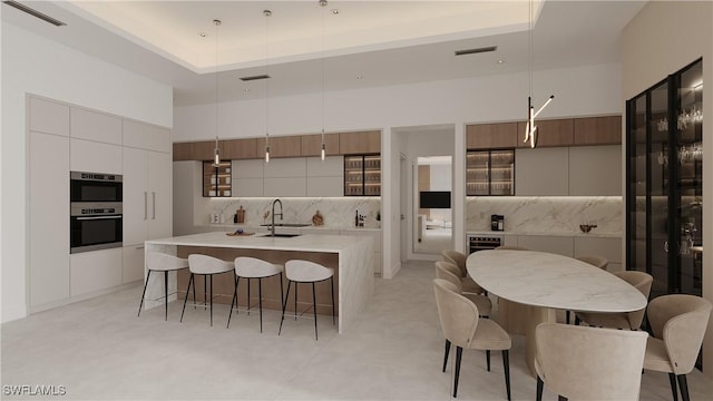 kitchen with backsplash, stainless steel double oven, a kitchen island with sink, sink, and pendant lighting