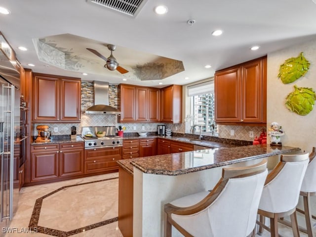 kitchen featuring kitchen peninsula, a raised ceiling, dark stone countertops, and wall chimney range hood