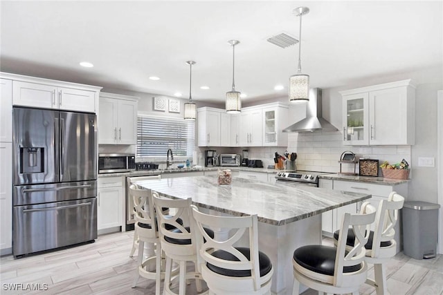 kitchen featuring a center island, wall chimney range hood, decorative light fixtures, white cabinetry, and stainless steel appliances