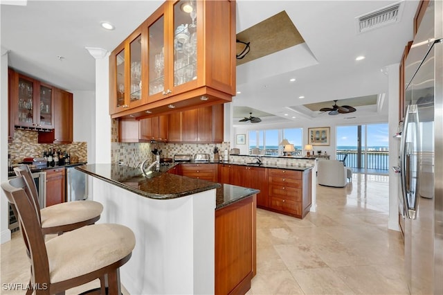 kitchen with kitchen peninsula, a kitchen breakfast bar, a tray ceiling, a water view, and dark stone countertops