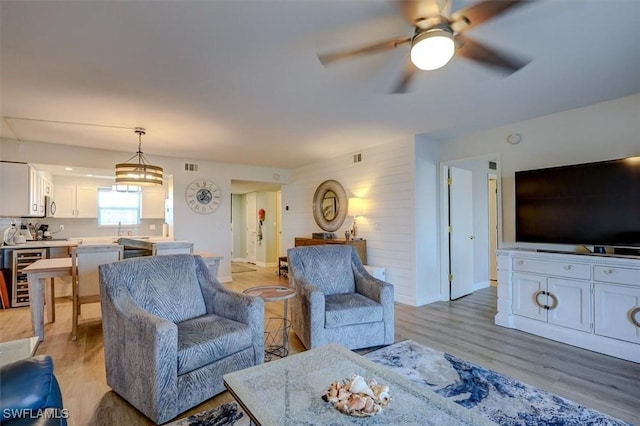 living room with wine cooler, wood walls, light hardwood / wood-style flooring, and ceiling fan with notable chandelier