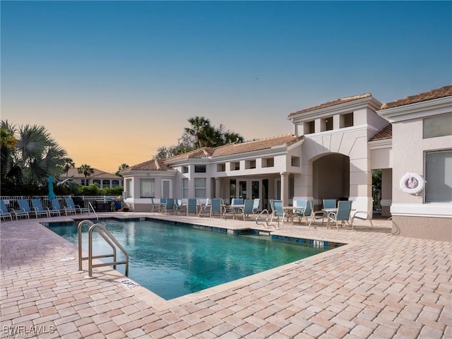 pool at dusk featuring a patio area, fence, and a community pool