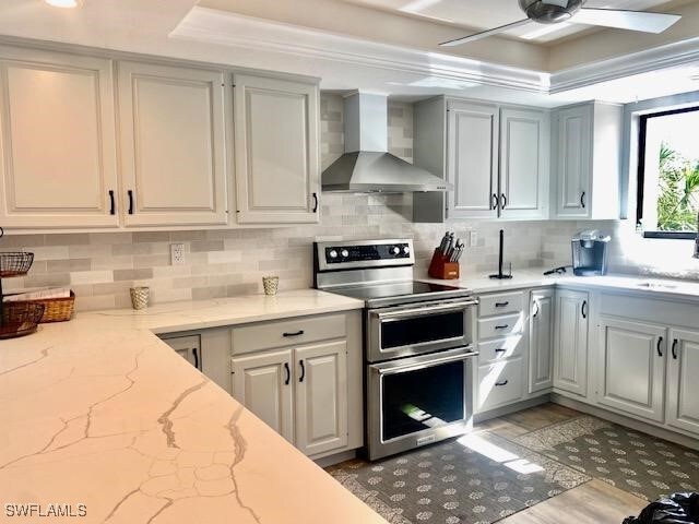 kitchen with light stone counters, wall chimney range hood, gray cabinets, and range with two ovens