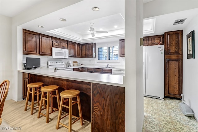 kitchen featuring ceiling fan, a breakfast bar area, kitchen peninsula, white appliances, and a tray ceiling