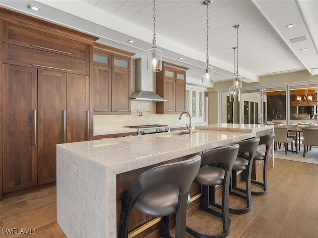 kitchen featuring a large island with sink, wall chimney exhaust hood, light stone counters, decorative light fixtures, and a kitchen bar