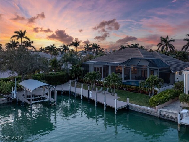 back house at dusk with a water view, glass enclosure, and a lawn