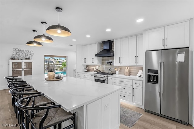 kitchen with appliances with stainless steel finishes, white cabinetry, hanging light fixtures, and wall chimney exhaust hood