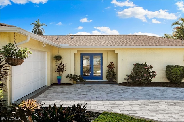 entrance to property featuring a garage, a shingled roof, brick siding, french doors, and stucco siding