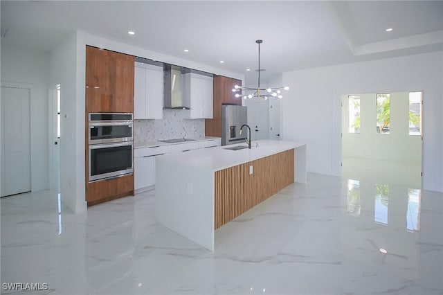 kitchen featuring tasteful backsplash, a kitchen island with sink, wall chimney range hood, decorative light fixtures, and white cabinets