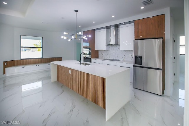 kitchen featuring wall chimney exhaust hood, an island with sink, appliances with stainless steel finishes, decorative light fixtures, and white cabinetry