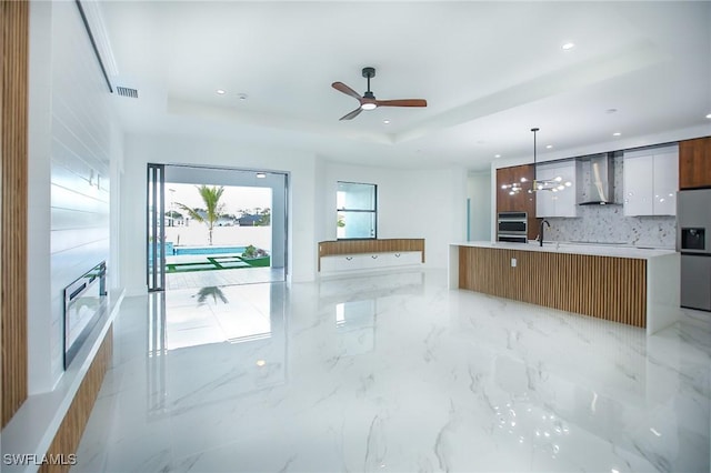 kitchen with a raised ceiling, a spacious island, wall chimney range hood, decorative light fixtures, and white cabinetry