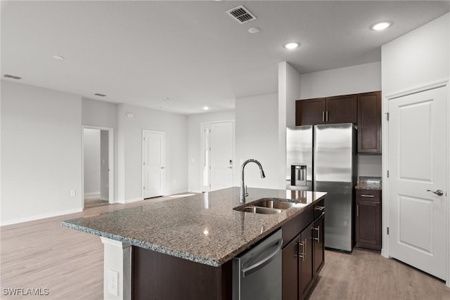 kitchen with a kitchen island with sink, sink, dark stone countertops, light wood-type flooring, and appliances with stainless steel finishes