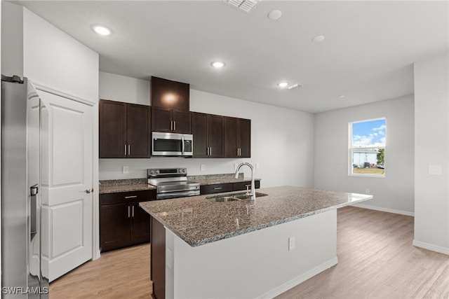 kitchen with sink, light stone counters, an island with sink, light hardwood / wood-style floors, and appliances with stainless steel finishes