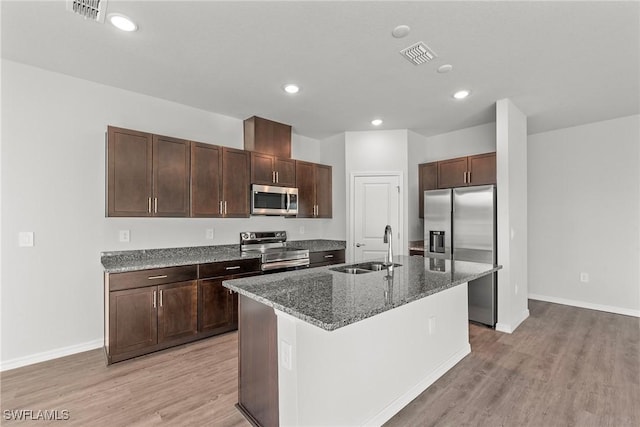 kitchen with appliances with stainless steel finishes, sink, a center island with sink, dark stone countertops, and light hardwood / wood-style floors