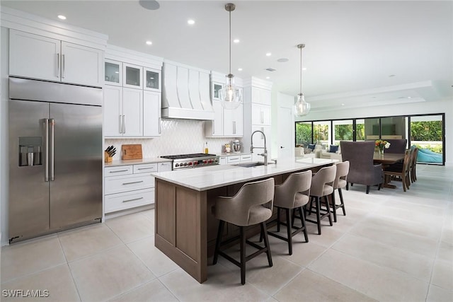 kitchen with sink, premium range hood, decorative light fixtures, a kitchen island with sink, and stainless steel built in fridge