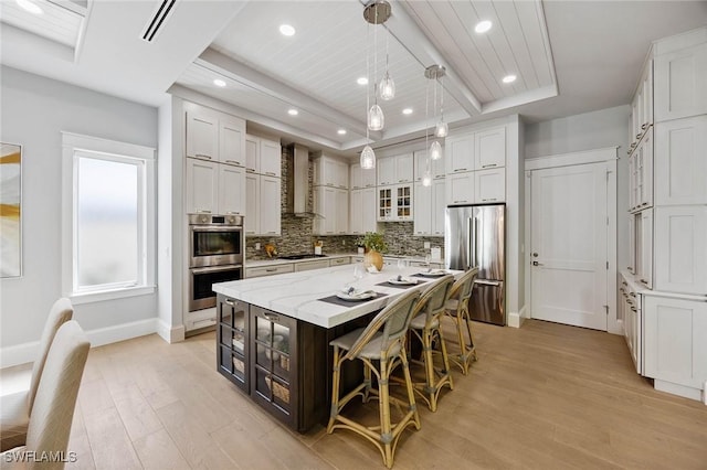 kitchen featuring light wood finished floors, a large island, appliances with stainless steel finishes, wall chimney range hood, and backsplash