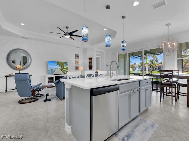 kitchen with a raised ceiling, hanging light fixtures, an island with sink, and stainless steel dishwasher