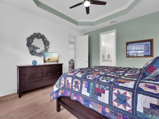 bedroom featuring light wood-type flooring, ensuite bathroom, a tray ceiling, ceiling fan, and crown molding