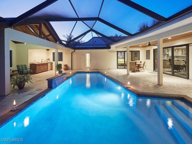 pool at dusk with a patio, glass enclosure, ceiling fan, and exterior kitchen