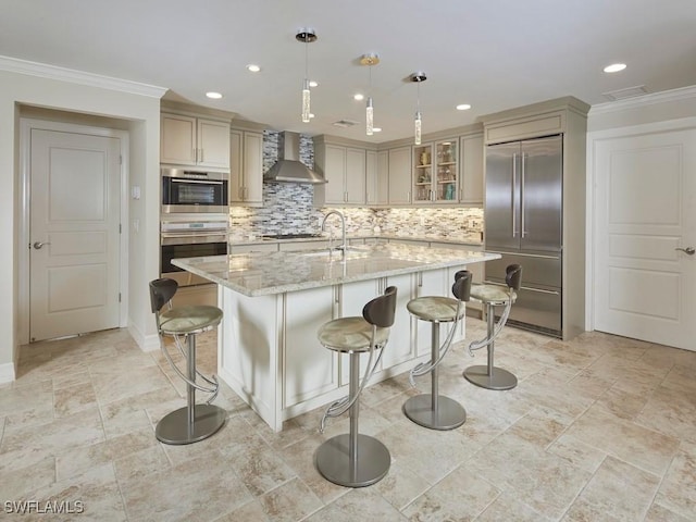 kitchen with wall chimney range hood, appliances with stainless steel finishes, a kitchen island with sink, hanging light fixtures, and light stone counters