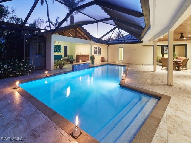 pool at dusk with glass enclosure, a patio area, and ceiling fan