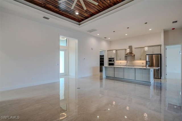 kitchen featuring wall chimney exhaust hood, stainless steel appliances, a kitchen island with sink, decorative light fixtures, and gray cabinets