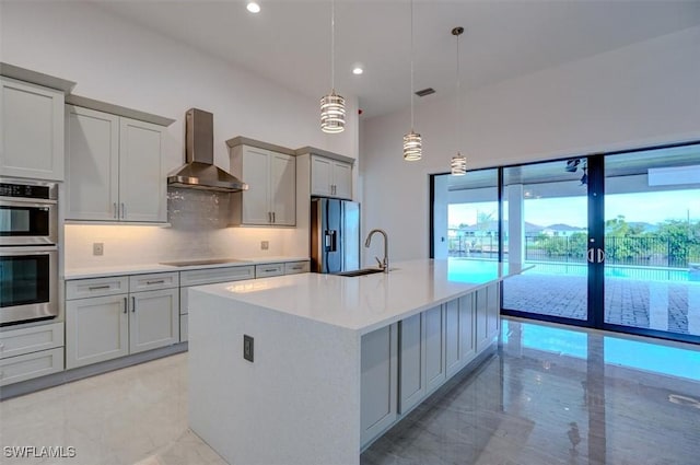 kitchen featuring appliances with stainless steel finishes, a kitchen island with sink, wall chimney range hood, decorative light fixtures, and gray cabinets