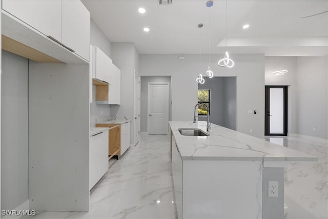 kitchen with a center island with sink, sink, hanging light fixtures, light stone counters, and white cabinetry