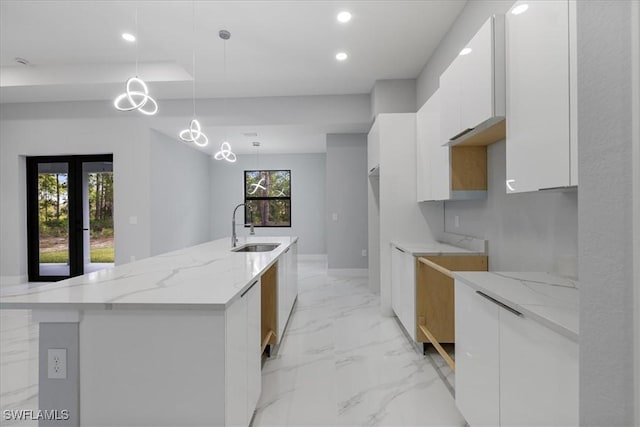 kitchen with a kitchen island with sink, white cabinets, sink, light stone countertops, and decorative light fixtures