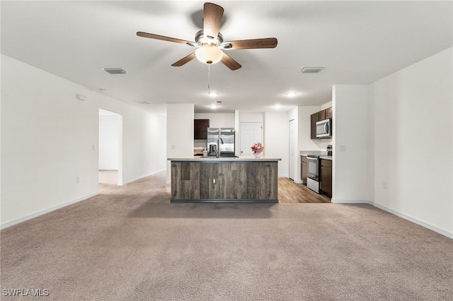 kitchen with light carpet, stainless steel appliances, an island with sink, ceiling fan, and dark brown cabinets