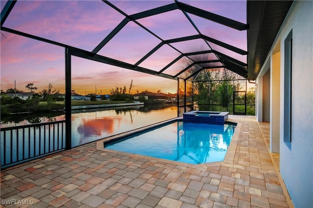 pool at dusk with an in ground hot tub, a water view, glass enclosure, and a patio area