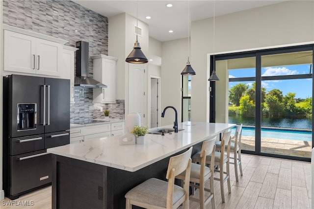 kitchen featuring wall chimney exhaust hood, hanging light fixtures, high quality fridge, a kitchen island with sink, and white cabinets