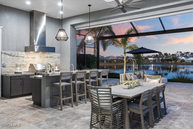 patio terrace at dusk with area for grilling, ceiling fan, a bar, a water view, and a grill