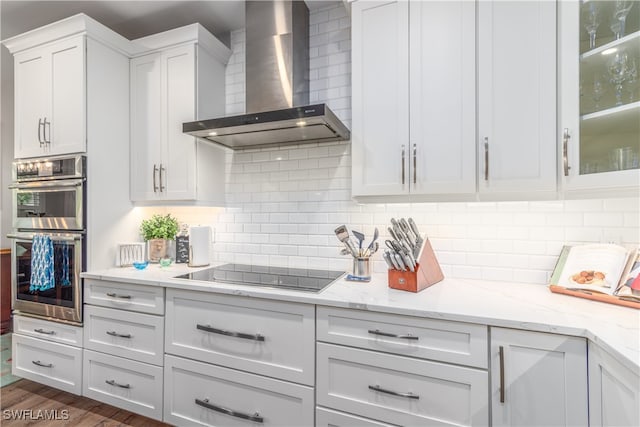 kitchen featuring white cabinets, black electric stovetop, stainless steel double oven, wall chimney range hood, and backsplash