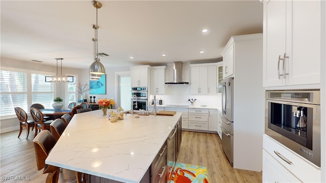 kitchen with light wood finished floors, wall chimney exhaust hood, a kitchen island with sink, stainless steel appliances, and white cabinetry