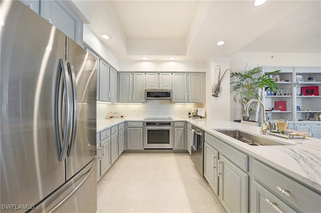 kitchen featuring sink, gray cabinets, appliances with stainless steel finishes, a tray ceiling, and light stone counters